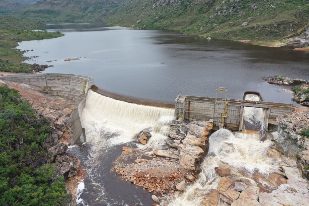 Barragem de Pequena Central Hidrelétrica (PCH) em área montanhosa, com reservatório cheio e água vertendo pelo vertedouro de concreto sobre rochas naturais.​