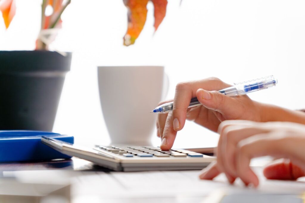 Mãos utilizando calculadora e caneta, com papéis sobre a mesa, representando atividades de análise financeira e organização de despesas.