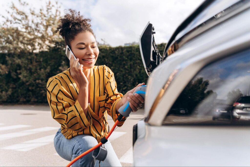 Mulher de cabelo preso e blusa amarela listrada conecta cabo de carregamento elétrico em carro branco, enquanto fala ao telefone em um estacionamento ao ar livre.