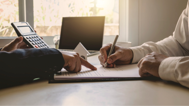 Duas pessoas em reunião à mesa analisando documentos financeiros, com uma delas apontando para uma planilha e outra anotando, enquanto uma calculadora aparece ao lado.